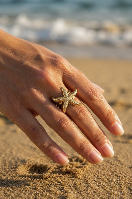 Bague étoile de mer dorée portée sur la main en bord de mer – grande étoile sculptée texturée sur fond de sable et vagues, acier inoxydable ajustable – Bijoux Incandescence