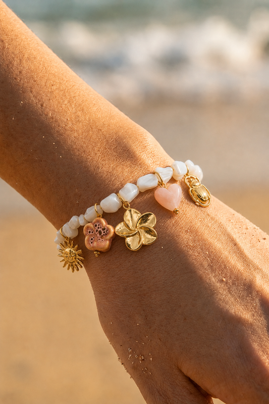 Bracelet charms bohème porté au poignet sur fond de plage — perles blanches, breloques dorées soleil, fleur, cœur et scarabée en acier inoxydable — Bijoux Incandescence