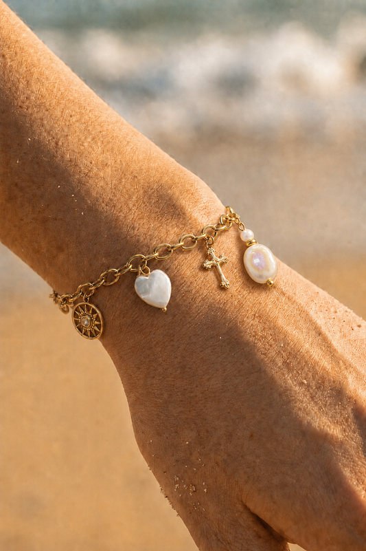 Bracelet charms doré porté au poignet sur fond de plage — médaillon soleil, cœur nacré, croix dorée et perle baroque en acier inoxydable — Bijoux Incandescence