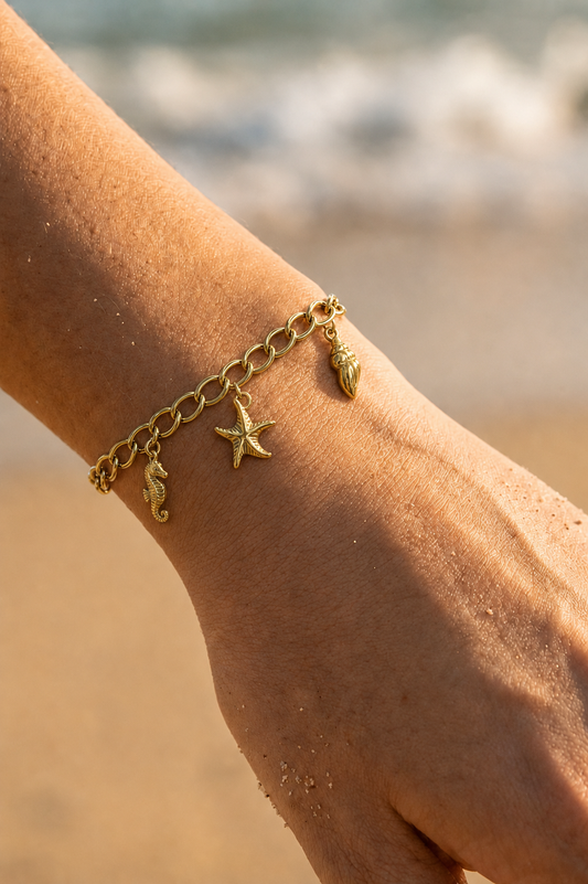 Bracelet charms doré porté au poignet sur fond de plage — hippocampe, étoile de mer et coquille sur chaîne gourmette en acier inoxydable — Bijoux Incandescence