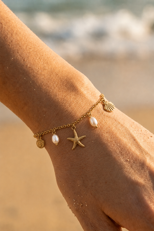 Bracelet charms doré porté au poignet sur fond de plage — étoile de mer, coquille Saint-Jacques, médaillon soleil et perles d'eau douce — acier inoxydable – Bijoux Incandescence