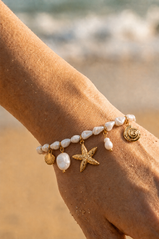 Bracelet charms doré porté au poignet sur fond de plage — perles baroques blanches, étoile de mer, coquille Saint-Jacques et spirale dorée en acier inoxydable — Bijoux Incandescence