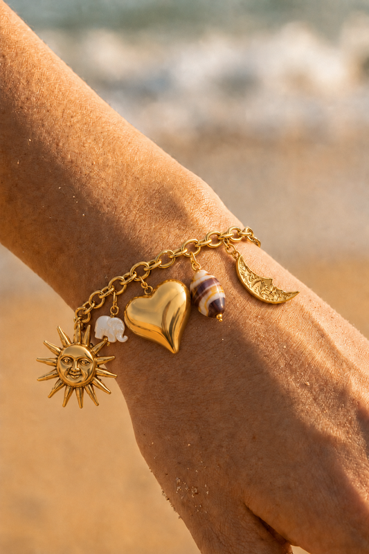 Bracelet charms doré porté au poignet sur fond de plage — soleil visage, cœur bombé, lune croissant, éléphant ivoire et perle rayée en acier inoxydable — Bijoux Incandescence