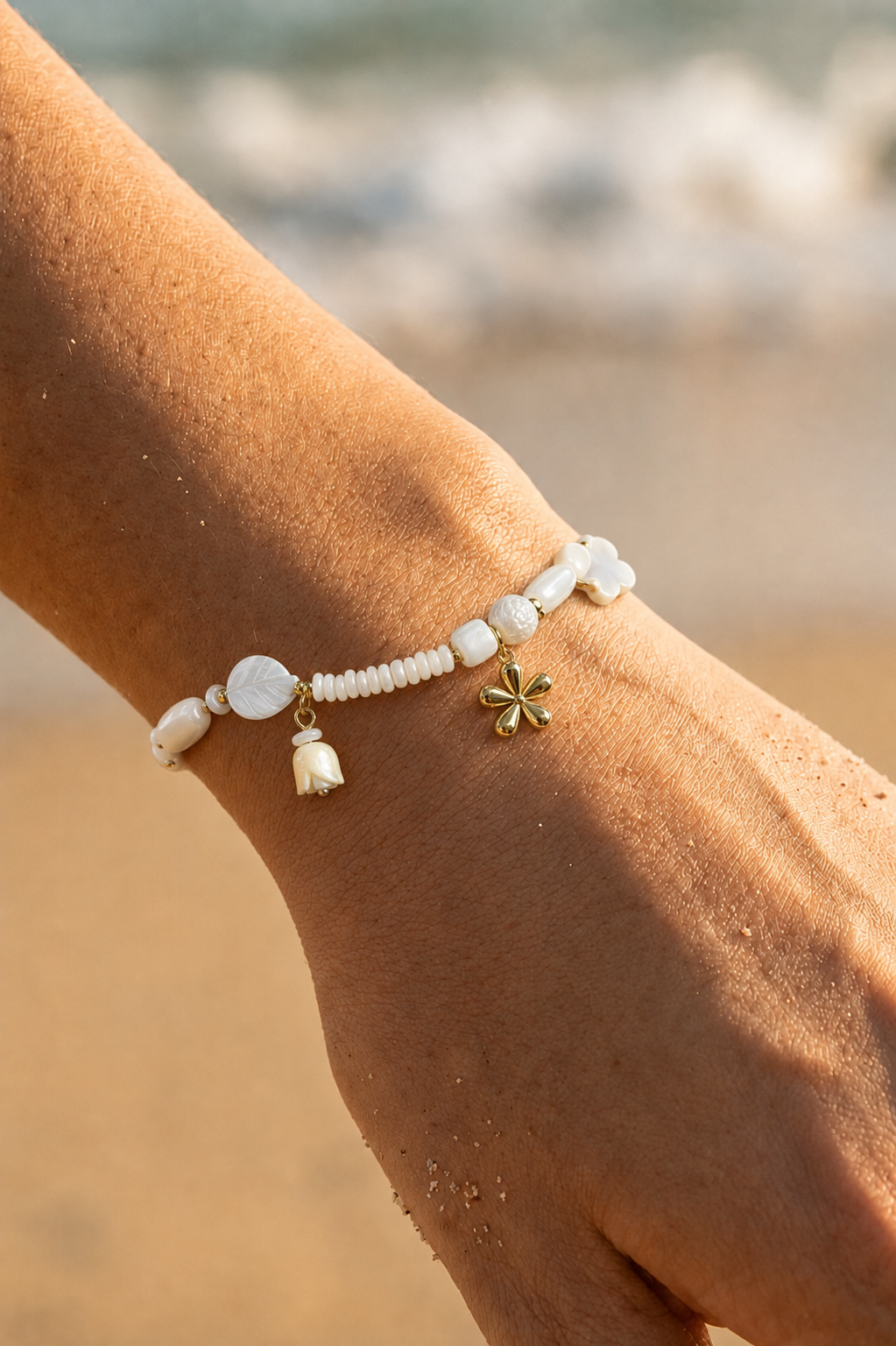 Bracelet charms porté au poignet sur fond de plage — perles nacrées blanches, tulipe nacrée et trèfle doré en acier inoxydable — Bijoux Incandescence