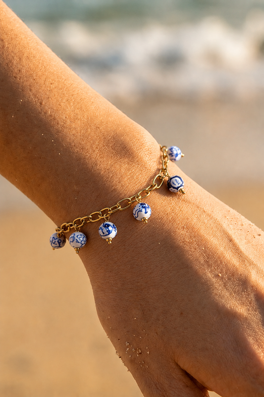 Bracelet charms doré porté au poignet sur fond de plage — perles rondes en porcelaine bleue et blanche style Delft sur chaîne dorée en acier inoxydable — Bijoux Incandescence