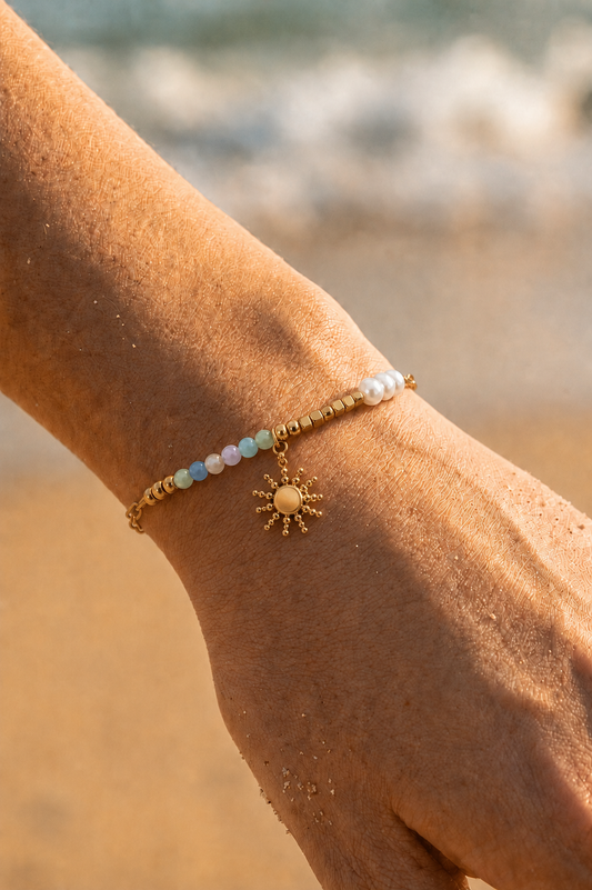 Bracelet charms porté au poignet sur fond de plage — perles pastel bleues et vertes, perles dorées et charm soleil doré en acier inoxydable — Bijoux Incandescence