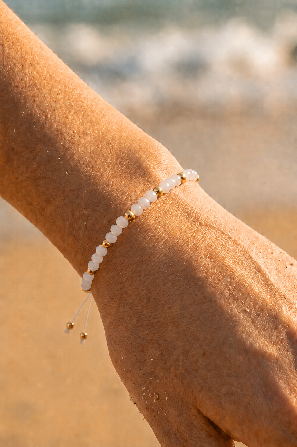 Bracelet fin cordon blanc ajustable porté au poignet sur fond de plage — petites perles nacrées blanches et perles dorées intercalées — Bijoux Incandescence