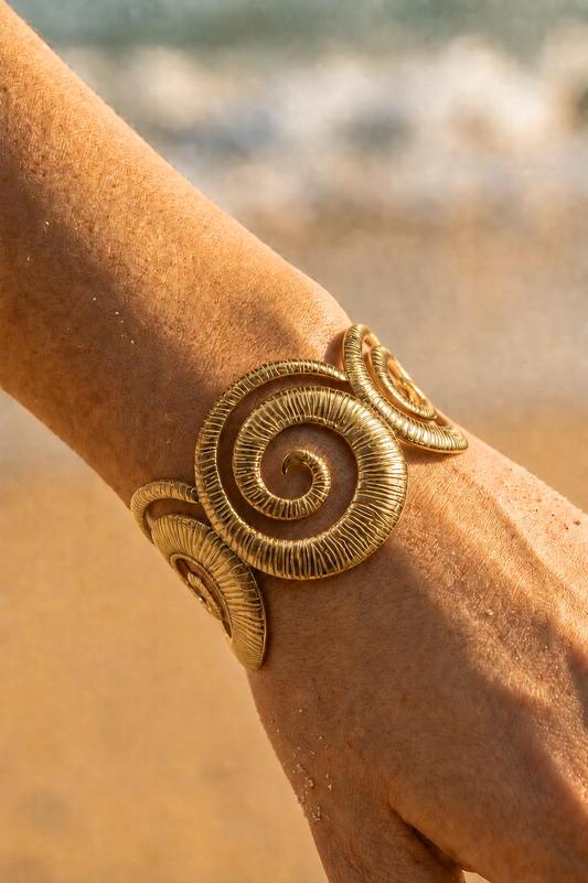 Bracelet jonc doré statement porté au poignet sur fond de plage — grandes spirales ammonite texturées en acier inoxydable — Bijoux Incandescence
