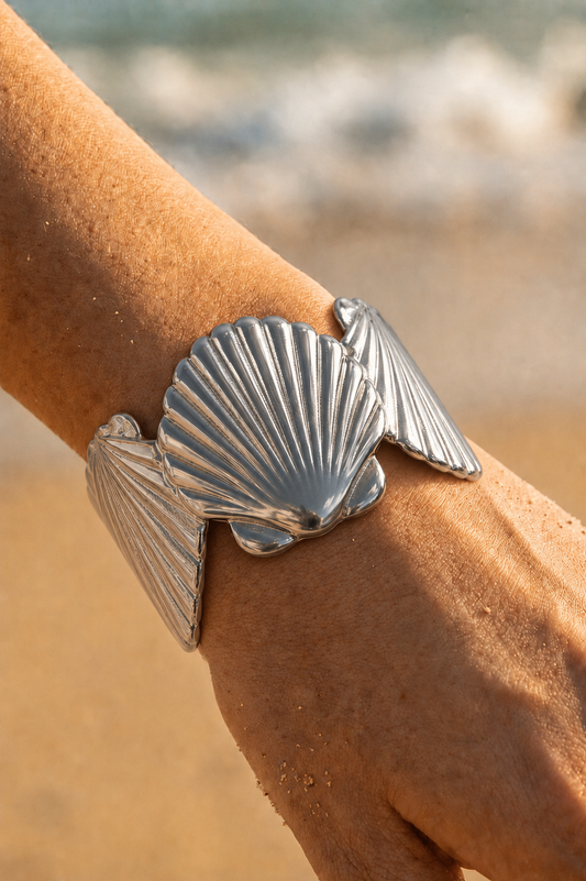 Bracelet jonc argenté porté au poignet sur fond de plage — grandes coquilles Saint-Jacques en relief en acier inoxydable — Bijoux Incandescence