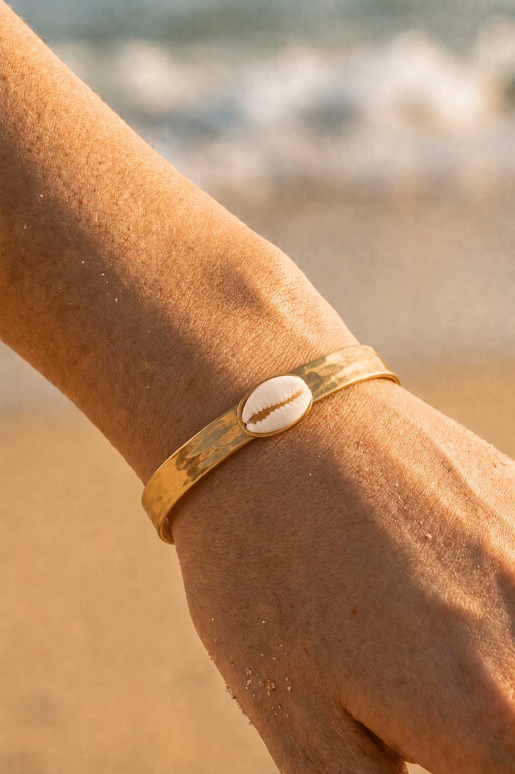 Bracelet jonc doré porté au poignet sur fond de plage — coquillage caurie blanc en acier inoxydable — Bijoux Incandescence