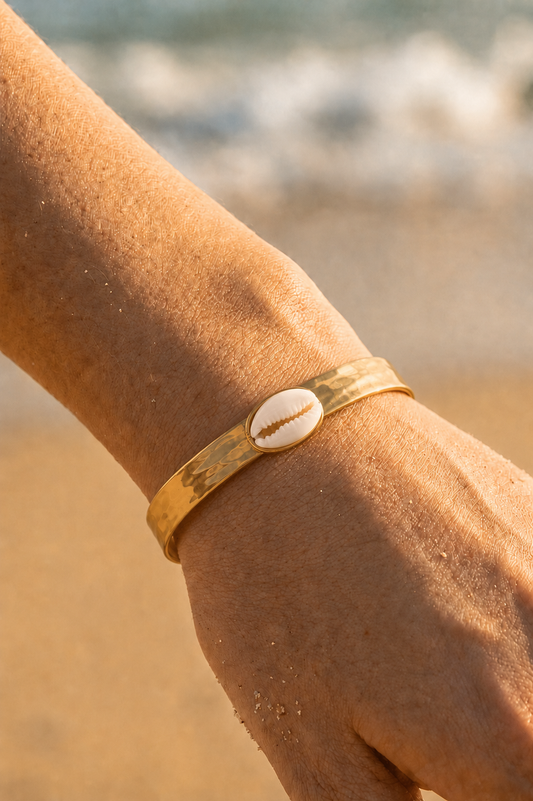 Bracelet jonc doré porté au poignet sur fond de plage — coquillage caurie blanc en acier inoxydable — Bijoux Incandescence