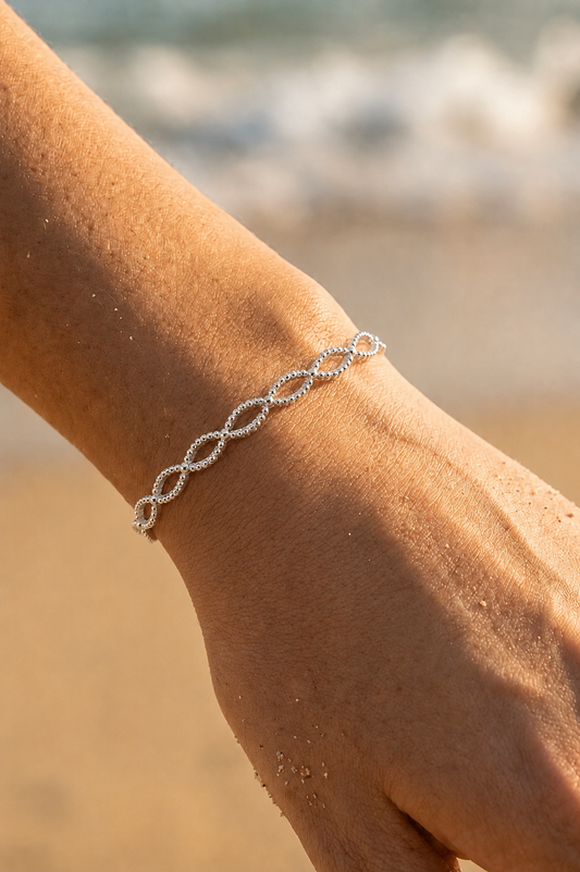 Bracelet jonc argenté tressé entrelacé porté au poignet sur fond de plage — motif infini en acier inoxydable — Bijoux Incandescence