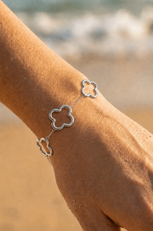Bracelet argenté porté au poignet sur fond de plage — trois trèfles ajourés sur fine chaîne en acier inoxydable — Bijoux Incandescence