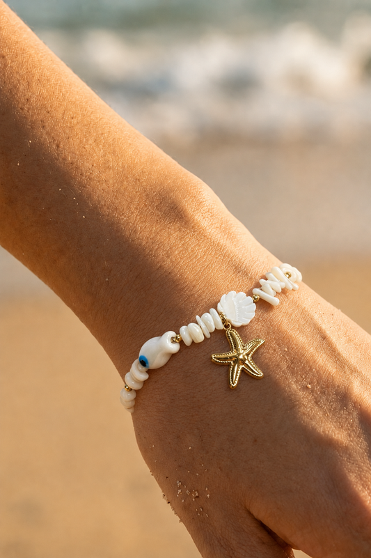 Bracelet trésor marin porté au poignet sur fond de plage — perles blanches, coquillage nacré, evil eye et étoile de mer dorée en acier inoxydable — Bijoux Incandescence